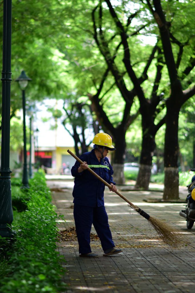 Urban maintenance worker sweeping a tree-lined path in a sunlit park, wearing safety gear.